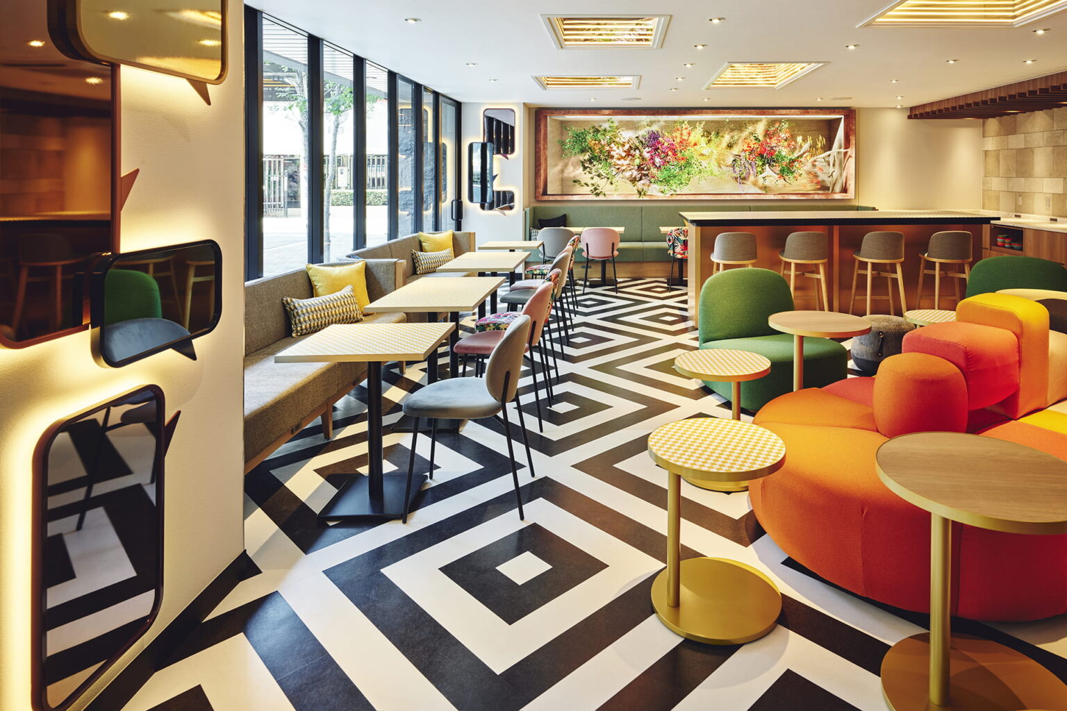 A colourful hotel lobby lounge with a black and white geometric floor, orange sofas and a floral wall installation.