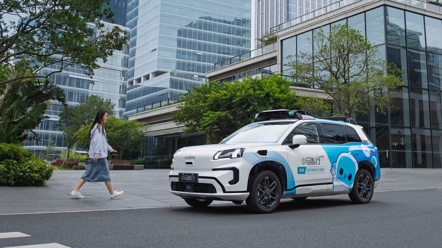 A white and blue Pony.ai robotaxi on a Shenzhen street as a pedestrian walks past glass office towers.